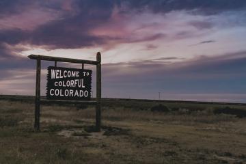 A wooden roadside sign reading "WELCOME TO COLORFUL COLORADO" standing in a grassy field against a twilight sky.