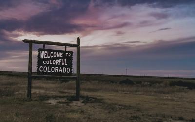 A wooden roadside sign reading "WELCOME TO COLORFUL COLORADO" standing in a grassy field against a twilight sky.
