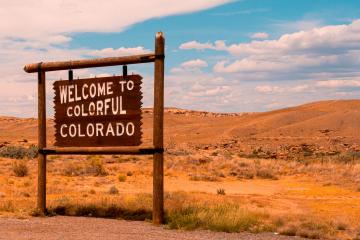 A rustic wooden sign reading "WELCOME TO COLORFUL COLORADO" mounted on a post, set against a backdrop of arid, rolling hills and a blue sky.