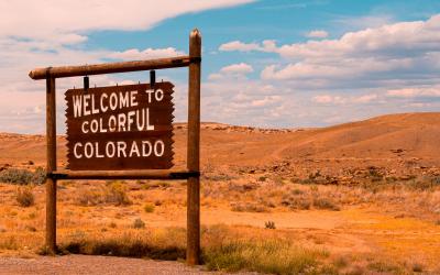 A rustic wooden sign reading "WELCOME TO COLORFUL COLORADO" mounted on a post, set against a backdrop of arid, rolling hills and a blue sky.