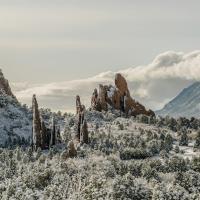 A winter landscape featuring distinctive red sandstone rock formations and snow-covered evergreen trees, with a larger mountain range in the background under a cloudy sky.