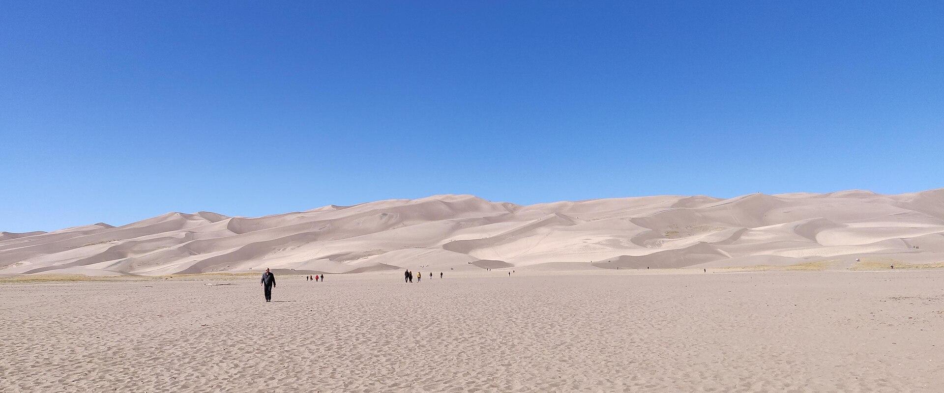 A wide landscape photograph showing vast sand dunes under a clear blue sky with several small figures of people walking on the sand.