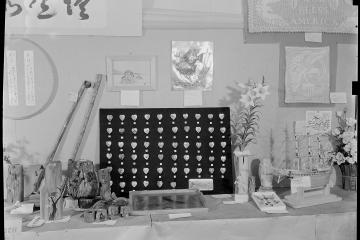 A black-and-white photograph of an indoor display table featuring various handmade crafts, including a model ship, wooden carvings, a heart-patterned board, and banners with Japanese calligraphy and "God Bless America."