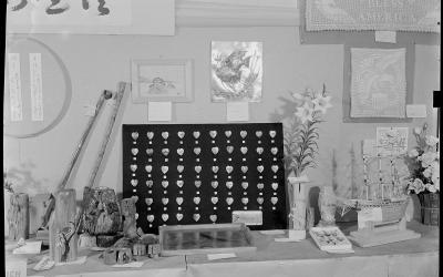 A black-and-white photograph of an indoor display table featuring various handmade crafts, including a model ship, wooden carvings, a heart-patterned board, and banners with Japanese calligraphy and "God Bless America."