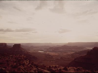 A vintage color photograph showing a wide canyon landscape with a river winding through the bottom, viewed from a high vantage point with rocky terrain in the foreground.