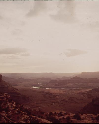 A vintage color photograph showing a wide canyon landscape with a river winding through the bottom, viewed from a high vantage point with rocky terrain in the foreground.