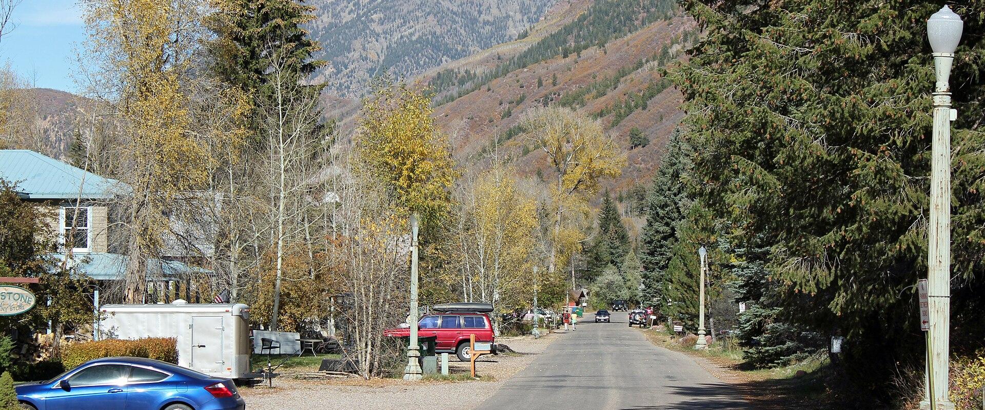 A paved road in a mountain town setting with autumn foliage, a blue car parked on the left, and a sign partially visible on the far left edge.