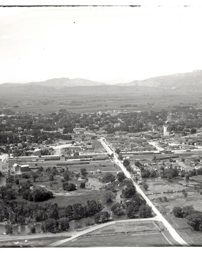 A vintage black-and-white aerial photograph showing a town situated along a winding river in the foreground, with a grid of streets and buildings extending toward a range of mountains in the background.