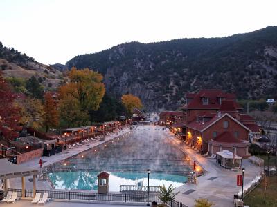 A high-angle view of a large, rectangular outdoor swimming pool with steam rising from the water, flanked by lounge chairs and a distinctive red brick building with a red roof, set against a backdrop of steep, forested hills.