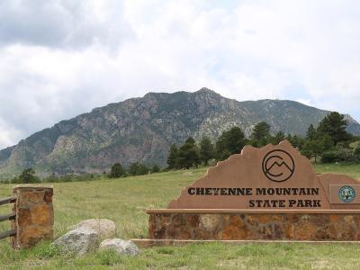 A large, custom-shaped brown sign with black lettering reading "CHEYENNE MOUNTAIN STATE PARK" and a Colorado state park logo, situated in a grassy field with a large mountain in the background.