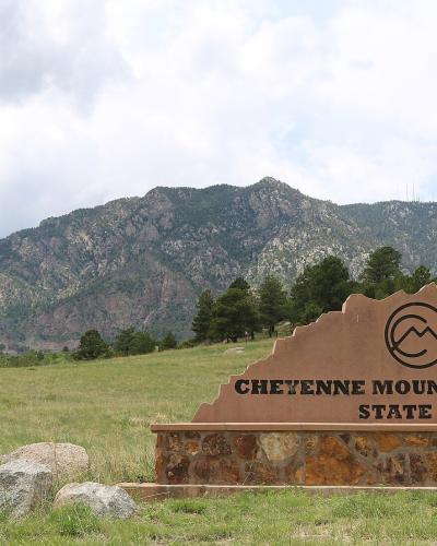 A large, custom-shaped brown sign with black lettering reading "CHEYENNE MOUNTAIN STATE PARK" and a Colorado state park logo, situated in a grassy field with a large mountain in the background.