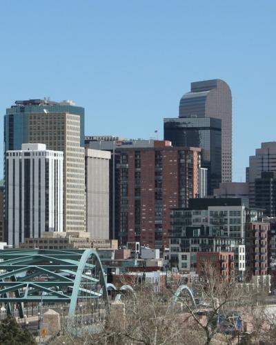 A city skyline featuring a distinctive teal-colored steel arch bridge in the foreground, with several skyscrapers behind it including one topped with a "Qwest" sign.