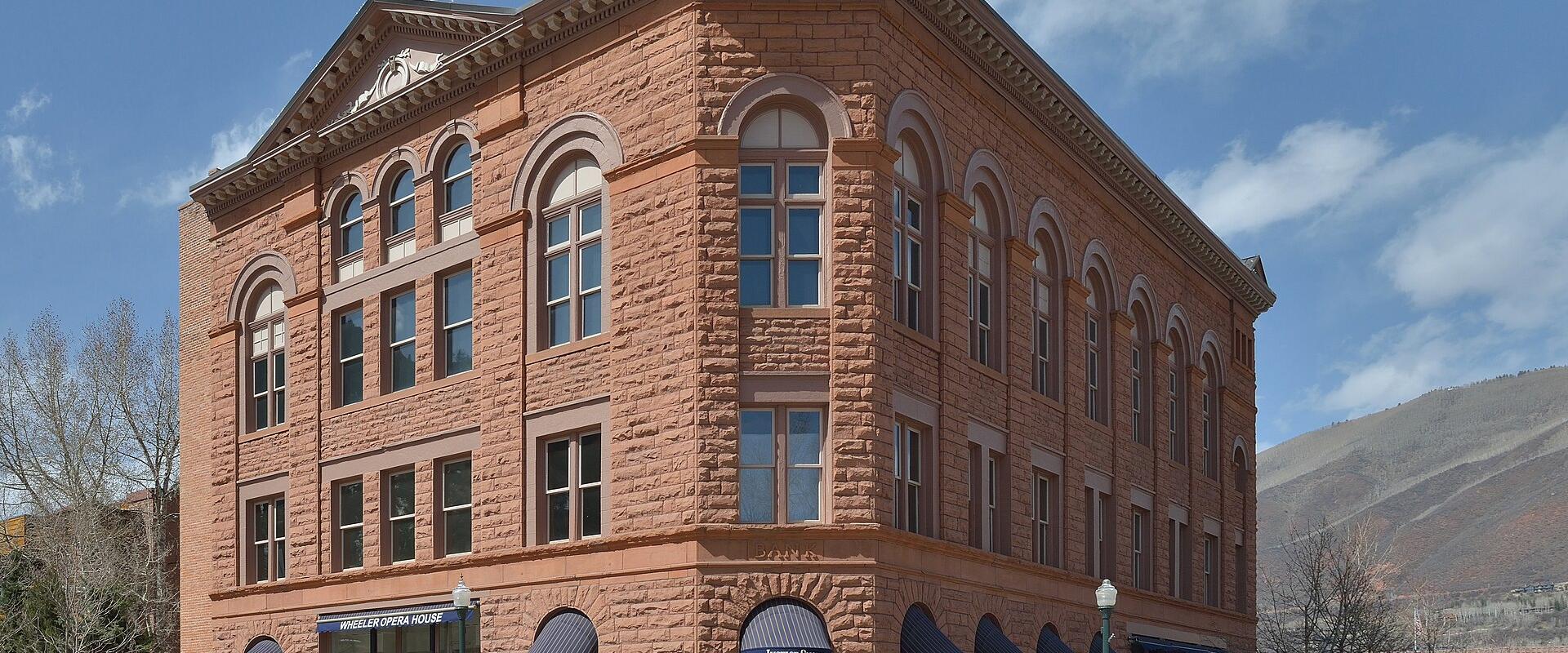 A three-story reddish-brown sandstone building on a street corner with blue awnings, featuring a sign above the entrance that reads "WHEELER OPERA HOUSE" and another sign for "Aspen's Shop".
