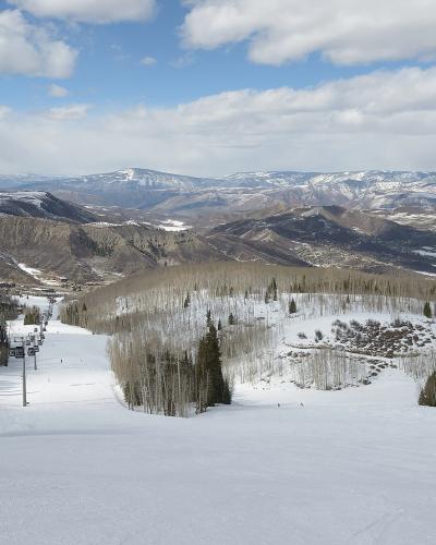 A view from a ski lift looking down a snowy slope with a skier, featuring black gondola cars suspended on cables against a backdrop of mountains and a blue sky.