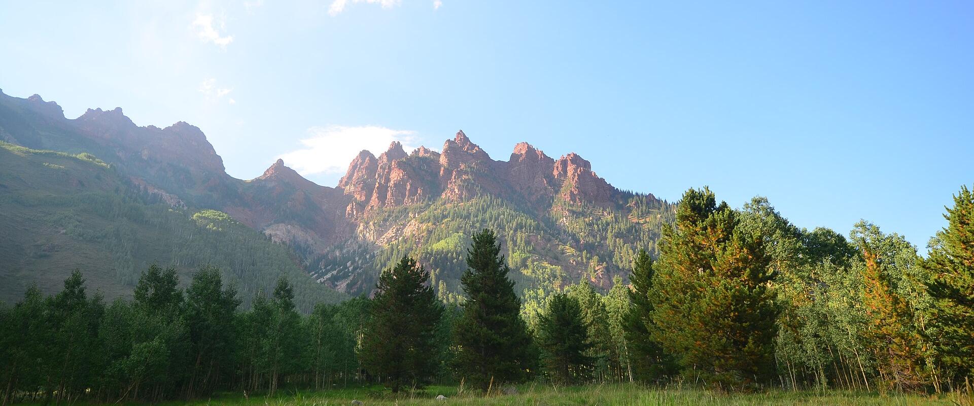 A landscape photograph showing a grassy meadow in the foreground, a line of coniferous and deciduous trees, and a prominent ridge of jagged, reddish-brown rock formations in the background under a blue sky.