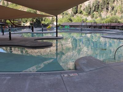 A large outdoor swimming complex featuring multiple interconnected pools, a water slide, and shade structures set against a steep, forested mountain backdrop.