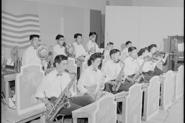 A black and white photograph showing a high school dance orchestra performing indoors, with musicians playing saxophones, trumpets, trombones, and violins behind music stands labeled "Music Makers".