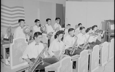 A black and white photograph showing a high school dance orchestra performing indoors, with musicians playing saxophones, trumpets, trombones, and violins behind music stands labeled "Music Makers".