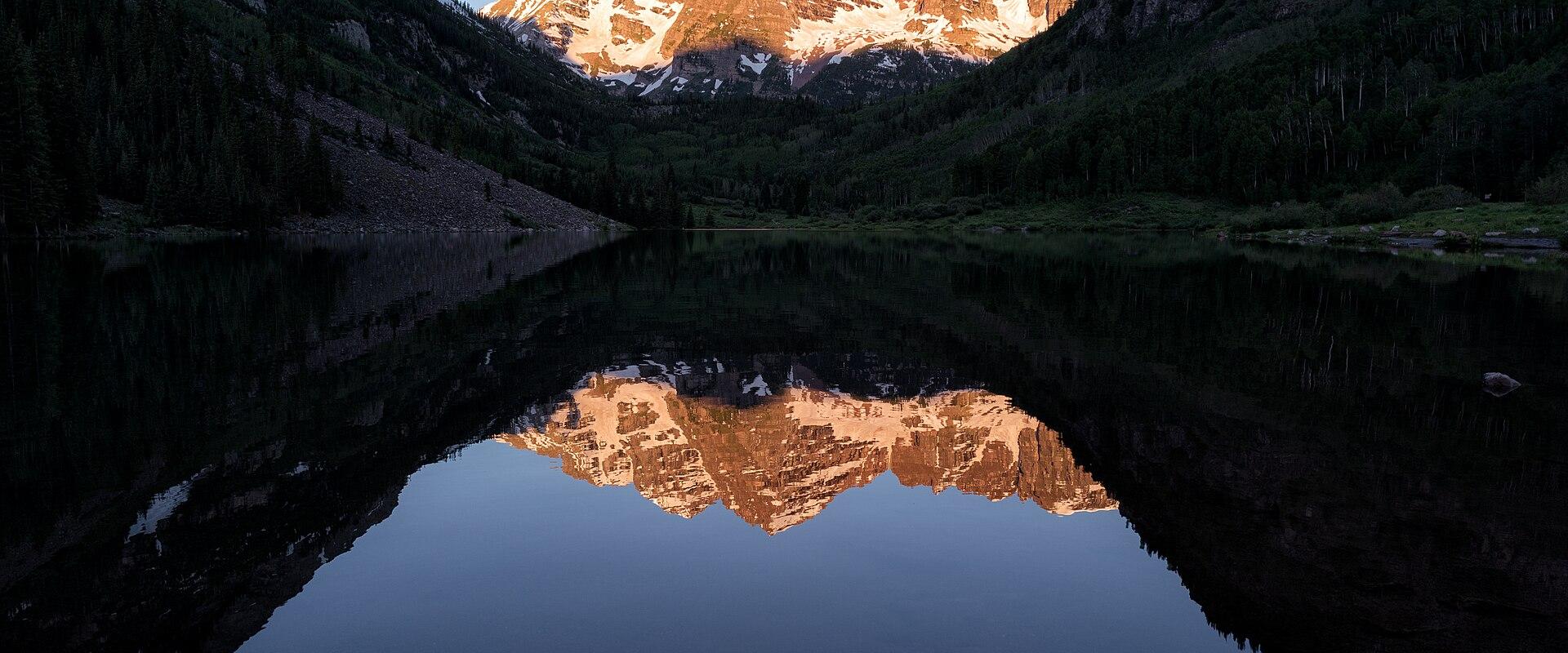 A landscape photograph showing two prominent, snow-dusted mountain peaks illuminated by sunlight, reflected in a calm lake in the foreground with forested slopes on either side.