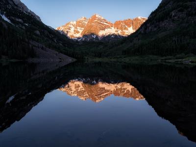 A landscape photograph showing two prominent, snow-dusted mountain peaks illuminated by sunlight, reflected in a calm lake in the foreground with forested slopes on either side.