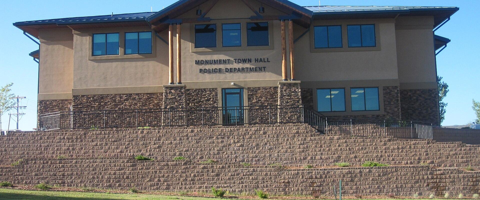 A two-story municipal building with a stone retaining wall in the foreground, featuring signage that reads "MONUMENT TOWN HALL POLICE DEPARTMENT".