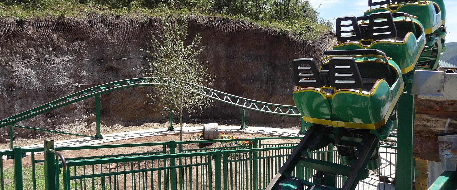 A green and yellow mine-cart style roller coaster train is shown on a track, with a rocky cliff face and trees in the background.