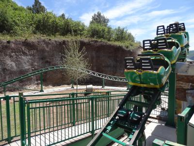 A green and yellow mine-cart style roller coaster train is shown on a track, with a rocky cliff face and trees in the background.