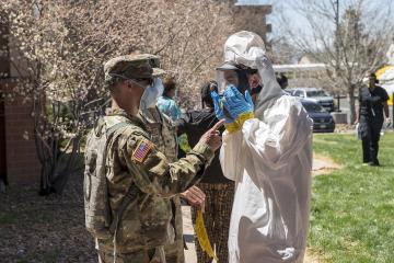 A US Army National Guard soldier in camouflage uniform and a person in a white hazmat suit with a face shield interact outdoors on a sunny day, with other people and vehicles in the background.