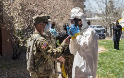 A US Army National Guard soldier in camouflage uniform and a person in a white hazmat suit with a face shield interact outdoors on a sunny day, with other people and vehicles in the background.