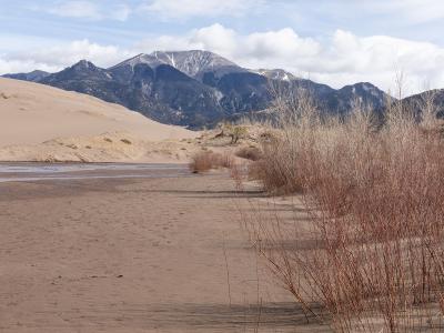 A landscape photograph showing a sandy riverbed with a stream, dry brush in the foreground, large sand dunes on the left, and a snow-capped mountain range in the background.