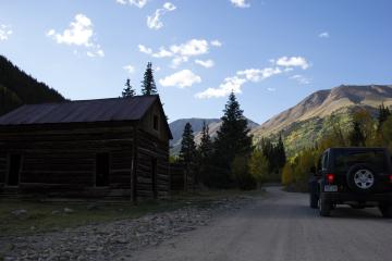 A weathered wooden cabin sits beside a dirt road with a black Jeep parked nearby, set against a backdrop of mountains and autumn-colored trees.