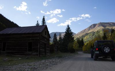 A weathered wooden cabin sits beside a dirt road with a black Jeep parked nearby, set against a backdrop of mountains and autumn-colored trees.