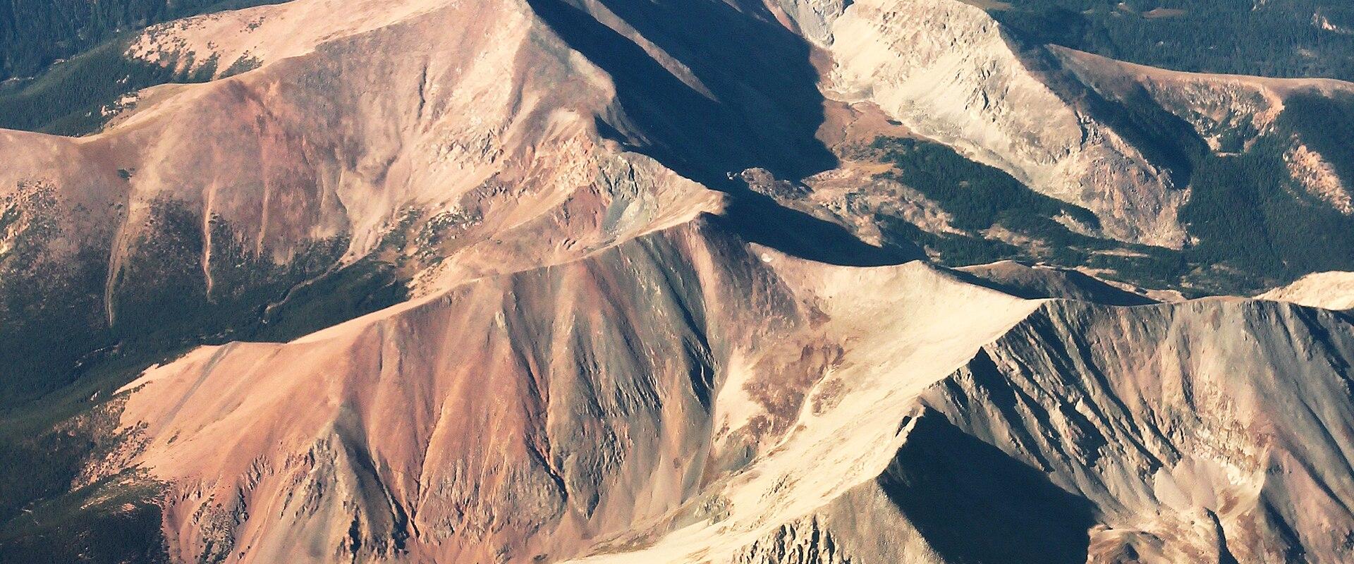 An aerial photograph showing rugged, multi-colored mountain ridges with exposed earth and rock formations, likely taken from an aircraft.