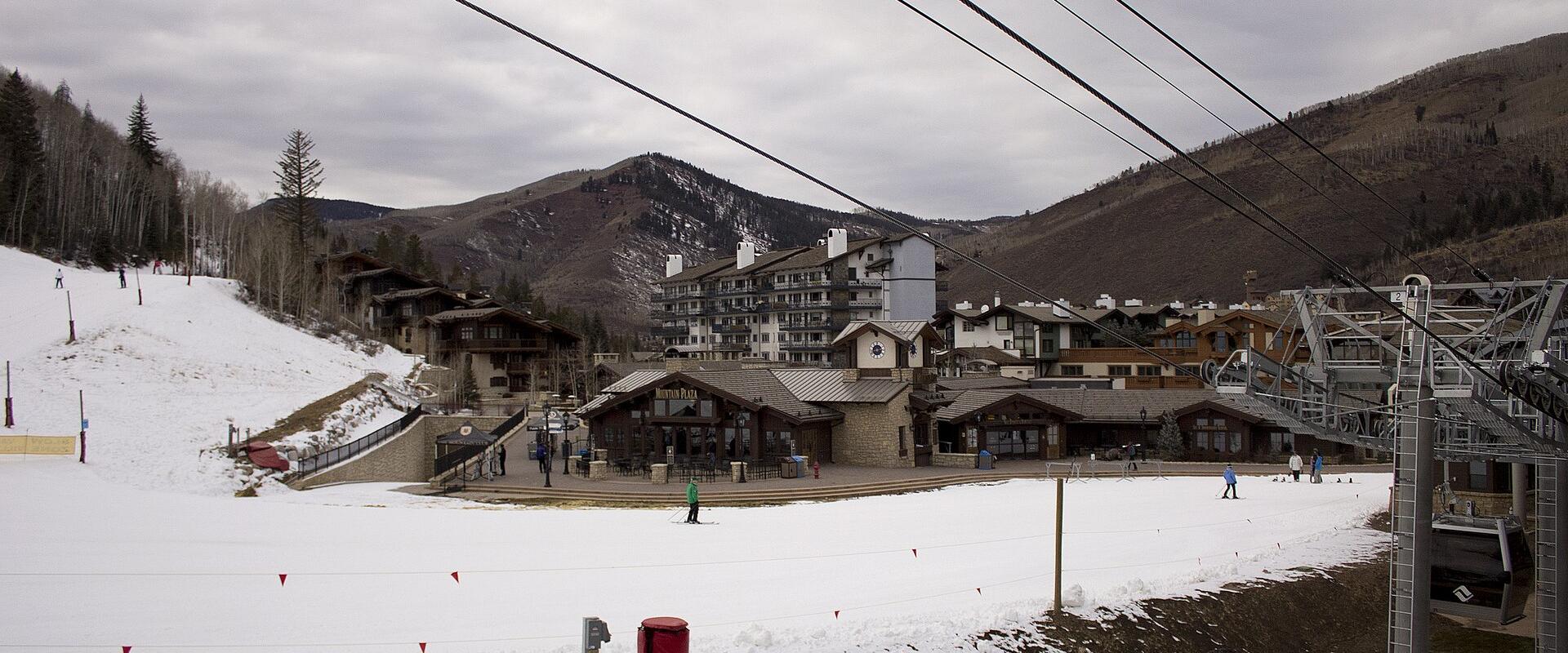 A snowy ski resort base area featuring a large multi-story hotel, several smaller lodge-style buildings, a chairlift structure on the right, and skiers on the snow under an overcast sky.
