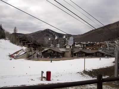A snowy ski resort base area featuring a large multi-story hotel, several smaller lodge-style buildings, a chairlift structure on the right, and skiers on the snow under an overcast sky.