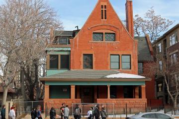 A red brick house with a steep gable and a group of people standing on the sidewalk in front of it.