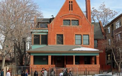 A red brick house with a steep gable and a group of people standing on the sidewalk in front of it.