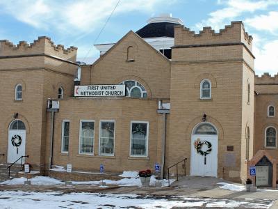 A tan brick church building with crenellated towers and a central cupola, featuring a prominent sign reading "FIRST UNITED METHODIST CHURCH" above the entrance.