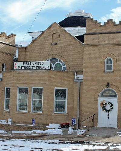 A tan brick church building with crenellated towers and a central cupola, featuring a prominent sign reading "FIRST UNITED METHODIST CHURCH" above the entrance.