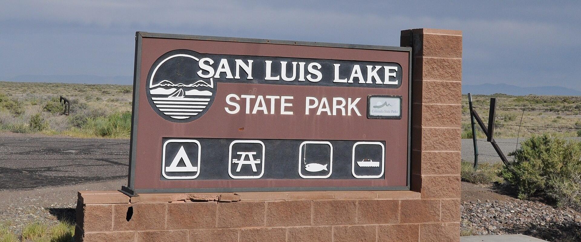 A large brown entrance sign for "SAN LUIS LAKE STATE PARK" mounted on a brick base, featuring icons for camping, picnicking, fishing, and boating, set against a high desert landscape.