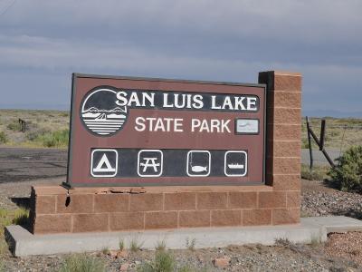 A large brown entrance sign for "SAN LUIS LAKE STATE PARK" mounted on a brick base, featuring icons for camping, picnicking, fishing, and boating, set against a high desert landscape.