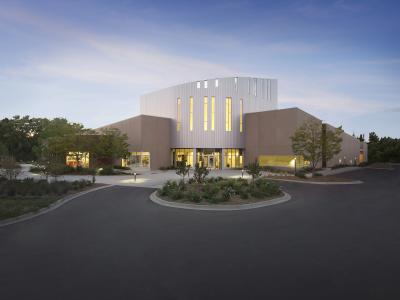 A modern, multi-winged institutional building with a central white vertical-slat facade and tan wings, illuminated from within at dusk, surrounded by a paved driveway and landscaping.