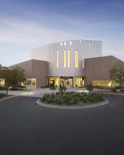 A modern, multi-winged institutional building with a central white vertical-slat facade and tan wings, illuminated from within at dusk, surrounded by a paved driveway and landscaping.