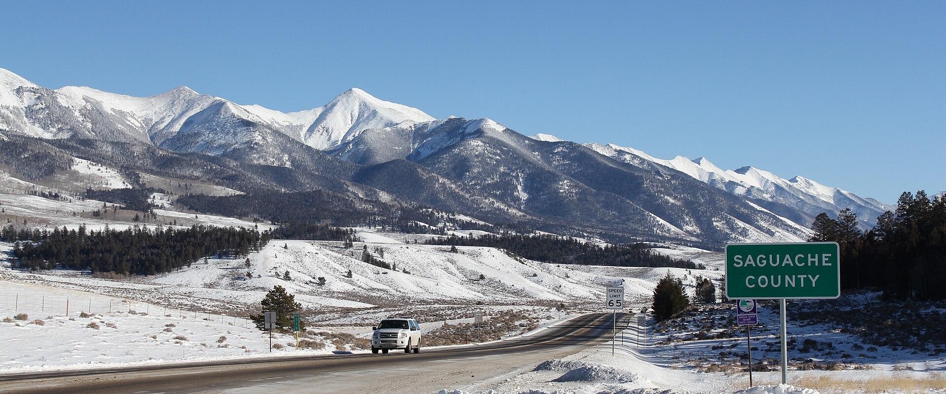 A snowy rural highway scene with a white pickup truck driving past a green road sign reading "SAGUACHE COUNTY" against a backdrop of snow-capped mountains.
