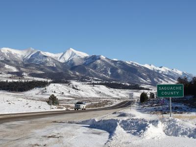A snowy rural highway scene with a white pickup truck driving past a green road sign reading "SAGUACHE COUNTY" against a backdrop of snow-capped mountains.