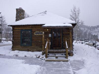 A snow-covered log cabin building with a stone chimney, wooden steps, and a sign reading "Breckenridge Heritage Alliance" mounted on the facade.