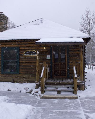 A snow-covered log cabin building with a stone chimney, wooden steps, and a sign reading "Breckenridge Heritage Alliance" mounted on the facade.