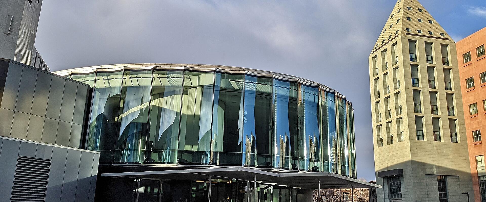 A low-angle exterior shot of a modern architectural complex featuring a curved glass facade and a tall, pyramid-topped stone tower under a cloudy sky.