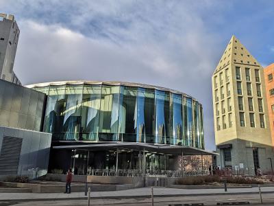 A low-angle exterior shot of a modern architectural complex featuring a curved glass facade and a tall, pyramid-topped stone tower under a cloudy sky.