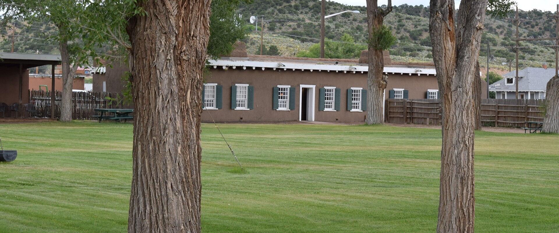 A long, single-story adobe-style building with white-trimmed windows and green shutters, viewed across a grassy lawn with large trees in the foreground.